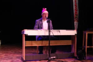 Występ muzyczno-wokalny Rafała Ugarenko. Dorosły mężczyzna w okularach, szarej marynarce i czapce Mikołaja siedzi na scenie i gra na białym keyboardzie. Instrument jest centralnym punktem, z mikrofonem zamontowanym na statywie przed muzykiem. Patrzy on przed siebie. Scena ma drewnianą podłogę i czarne tło. Z prawej strony instrumentu widoczny jest czarny zwinięty kabel, a za nim stoi drewniany stołek. Z lewej strony, w tle, widoczny jest fragment dekoracji.