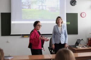 Zdjęcie z sali wykładowej, ujęcie zza ławek. Dwie wykładowczynie: Anna Bieganowska-Skóra w różowym kardiganie po lewej patrzy i gestykuluje w stronę widowni. Dorota Krać w jasnoniebieskim żakiecie stoi po prawej, skierowana w stronę audytorium. W tle na rzutniku wyświetlany jest kadr krajobrazowy (trawa, drzewa, drewniane sprzęty nad wodą).
