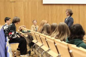 Interakcja między prowadzącymi a uczestnikami w auli wykładowej. Po prawej stronie kadru stoi dr hab. Andrzej Kapusta, zwrócony profilem do słuchaczy. W centrum, za ławami, siedzi uśmiechnięta prof. Halina Rarot. Na pierwszym planie i po lewej stronie widoczne są sylwetki studentów siedzących w rzędach jasnych, drewnianych ław oraz na niebieskich krzesłach.