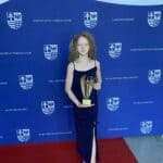 Na ciemnoniebieskim tle z powtarzającymi się cyklicznie białymi: herbem Województwa Świętokrzyskiego i napisem "Województwo Świętokrzyskie" stoi szczupła dziewczynka z kręconymi blond włosami do ramion w rękach trzyma statuetkę Świętokrzyski Anioł Dobroci w kolorze złotym