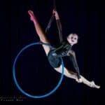 Dziewczynka ubrana w czarny połyskliwy kostium wykonuje akrobacje na niebieskiej obręczy, zawieszonej w powietrzu na czarnej szarfie. Tło fotografii jest czarne. Fot. Malowanie Światłem Tomasz Gas