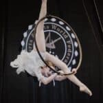Dziewczynka ubrana w kremowy kostium wykonuje akrobacje na obręczy, zawieszonej w powietrzu.