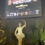 Dziewczyna w jasnym stroju akrobatycznym pozuje na tle czarnej kurtyny, u góry na ekranie wyświetlone są dane Mistrzostw Świata "Pole and Aerial World Cup" BBologna, November 27/28/29/30, 2025