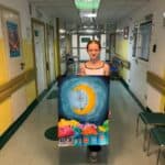 Korytarz placówki medycznej. Na środku stoi dziewczynka, trzyma przed sobą namalowany obraz przedstawiający księżyc ponad dachami budynków