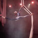 Dziewczynka ubrana w czarny kostium z kolorowymi wstawkami wykonuje akrobacje na podwójnej szarfie zawieszonej w powietrzu. Prawa stopa wyprostowanej nogi znajduje się z przodu sylwetki, pomiędzy splecionymi ze sobą szarfami, lewa wyprostowana noga jest wyciągnięta w tył - dziewczynka wykonuje w powietrzu szpagat