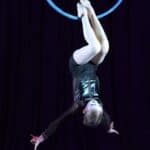 Dziewczynka ubrana w czarny połyskliwy kostium wykonuje akrobacje na czarnej obręczy, zawieszonej w powietrzu. Dziewczynka zwisa głową w dół, trzymając się obręczy jedynie stopami