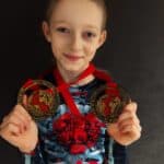 Dziewczynka pozuje do fotografii. W rękach trzyma zawieszone na szyi na czerwonych wstęgach dwa medale zdobyte podczas Mistrzostw Świata "Pole and Aerial World Cup". Uśmiecha się delikatnie