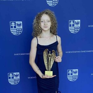 Na ciemnoniebieskim tle z powtarzającymi się cyklicznie białymi: herbem Województwa Świętokrzyskiego i napisem "Województwo Świętokrzyskie" stoi szczupła dziewczynka z kręconymi blond włosami do ramion w rękach trzyma statuetkę Świętokrzyski Anioł Dobroci w kolorze złotym