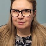 Portret długowłosej blondynki w czarnych okularach, delikatnie uśmiechniętej i patrzącej wprost przed siebie. Ma złote kolczyki oraz czarną górną część stroju w białe wzory.