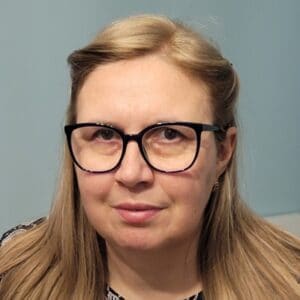 Portret długowłosej blondynki w czarnych okularach, delikatnie uśmiechniętej i patrzącej wprost przed siebie. Ma złote kolczyki w uszach.