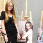 Fotografia autorstwa Agnieszki C. Gałązki. W centrum kadru przy sztaludze ze swoim zdjęciem, będącym częścią wystawy fotograficznej pt. „Czy mnie (nie) widzisz?” pozuje młoda kobieta. Kobieta ma jasne proste włosy, okulary i jest ubrana w czarną sukienkę