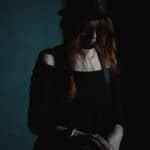 Rudowłosa kobieta ubrana na czarno pozuje w ciemnym pomieszczeniu, z czarnym kapeluszem na głowie. Spogląda w dół. Całą scenę utrzymano w przyciemnionej, nastrojowej tonacji