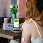 Fotografia autorstwa Pauliny Muszyńskiej, będąca częścią wystawy fotografii „Czy mnie (nie) widzisz?”. Rudowłosa kobieta siedzi tyłem przy małym stoliku, dotykając ekranu smartfona opartego o wazon z tulipanami. Na jej lewym przedramieniu widać okrągły sensor CGM do monitorowania glikemii. Na ekranie telefonu wyświetla się wykres i wartość „121” na zielonym tle. Przed nią stoi talerzyk z kawałkiem ciasta, a w tle dwa kubki kawy i ściana z graficznymi łukami w szarym i ceglastoczerwonym kolorze