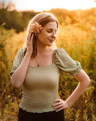 Rudowłosa kobieta pozuje w plenerze, opiera lewą rękę na biodrze, a prawą dotyka włosów przy prawym uchu. Spogląda w dół z delikatnym uśmiechem. W tle widać roślinność w ciepłych, jesiennych barwach.