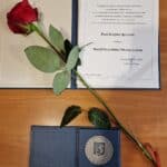 Na drewnianym blacie leżą: u góry dokument w granatowej, twardej oprawie o przyznaniu Medalu Prezydenta Miasta Lublin Justynie Spryszak, a poniżej srebrny, okrągły medal w granatowym pudełku. Przez oba przedmioty po skosie ułożona jest czerwona róża z zielonymi liśćmi i czerwoną wstążką.