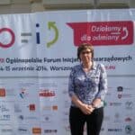 Krótkowłosa kobieta w okularach pozuje na tle ścianki z napisem „Ogólnopolskie Forum Inicjatyw Pozarządowych, 14–15 września 2014, Warszawa”, w dolnej części ścianki widoczne są liczne logotypy organizatorów, partnerów i patronów medialnych