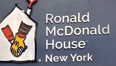Zbliżenie na szyld. Od lewej strony kształt domu z umieszczonymi wewnątrz splecionymi dłońmi, obok biały napis: "Ronald McDonald House New York" na szaroniebieskim tle