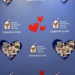 Na niebieskim tle planszy rozmieszczono grafiki mniejszych, czerwonych serc czerwonych oraz większych z kolażem zdjęć oraz napisy "Celebrating 45 Years" wraz z logotypem Ronald McDonald House New York