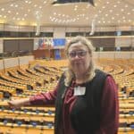 Długowłosa blondynka w okularach pozuje w sali obrad plenarnych Parlamentu Europejskiego w Brukseli, w charakterystycznej półokrągłej przestrzeni (2025 r.).