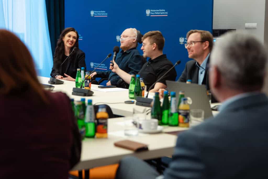 Sześć osób siedzi przy stole konferencyjnym z mikrofonami, notatnikami i napojami, uczestnicząc w dyskusji, na tle niebieskiego tła z oficjalnymi logotypami