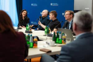 Sześć osób siedzi przy stole konferencyjnym z mikrofonami, notatnikami i napojami, uczestnicząc w dyskusji, na tle niebieskiego tła z oficjalnymi logotypami