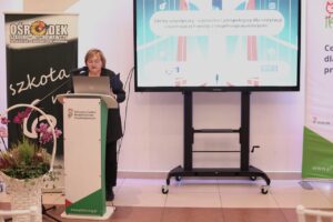 Kobieta w okularach przemawia z mównicy PFRON (Państwowy Fundusz Rehabilitacji Osób Niepełnosprawnych), czyta z laptopa. Na ekranie stojącym obok niej widoczny jest tytuł: "Mosty współpracy – wyzwania i perspektywy dla instytucji wspierających osoby z niepełnosprawnościami". W tle baner Ośrodka Szkolno-Wychowawczego