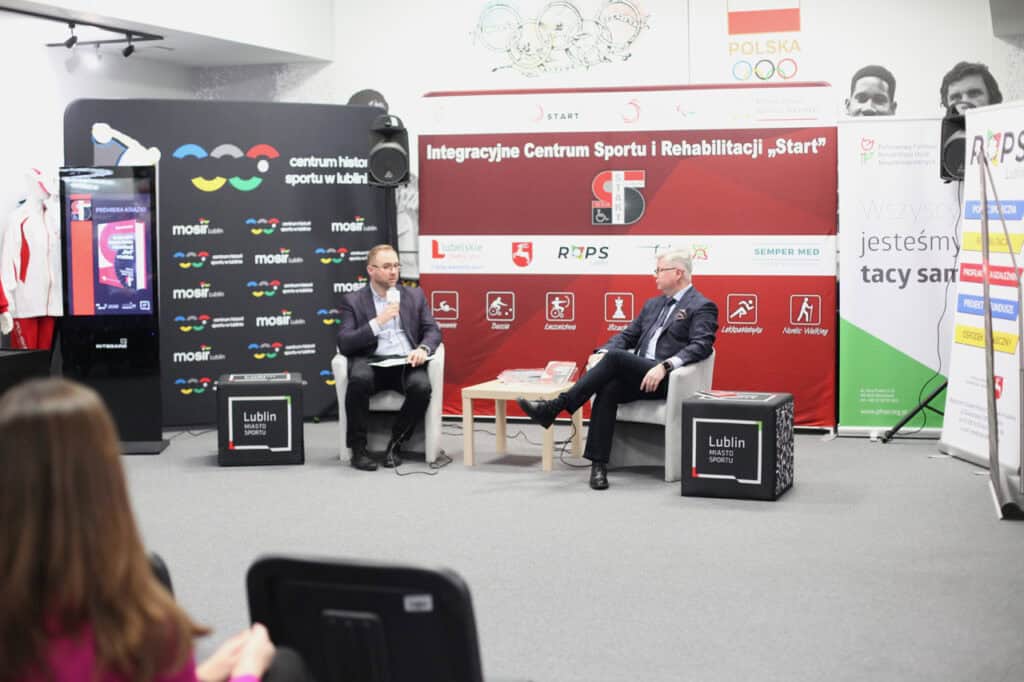 Dwóch elegancko ubranych mężczyzn (po prawej prof. Robert Litwiński) siedzi w fotelach podczas panelu dyskusyjnego. Rozmawiają przed dużym czerwono-czarnym banerem promującym „Integracyjne Centrum Sportu i Rehabilitacji 'Start'" z logotypami Lublina. Widok z widowni