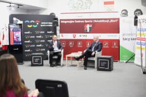 Dwóch elegancko ubranych mężczyzn (po prawej prof. Robert Litwiński) siedzi w fotelach podczas panelu dyskusyjnego. Rozmawiają przed dużym czerwono-czarnym banerem promującym „Integracyjne Centrum Sportu i Rehabilitacji 'Start'" z logotypami Lublina. Widok z widowni