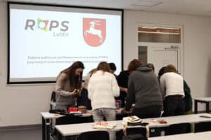 Grupa osób stojących przy białych stołach w nowoczesnej sali wykładowej, pochylonych nad wspólnym zadaniem warsztatowym. W tle, na ścianie, znajduje się duży ekran projekcyjny, na którym wyświetlone są logotypy Regionalnego Ośrodka Polityki Społecznej (ROPS) w Lublinie oraz herb województwa lubelskiego. Pod grafikami widnieje napis informujący, że zadanie publiczne jest finansowane ze środków PFRON przyznanych przez Samorząd Województwa Lubelskiego.