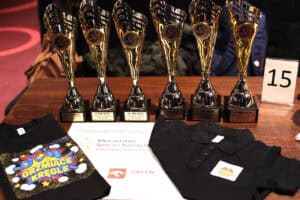 Sześć błyszczących, złotych pucharów z czarnymi podstawami (trofea za 1, 2 i 3 miejsce), ułożonych w rzędzie na drewnianym stole. Przed nimi leżą czarne koszulki (t-shirt z kolorowym napisem „GRZMIĄCE KRĘGLE", koszulka polo z logo) oraz białe pismo sponsorskie z logotypami: Fundacji ORLEN oraz Ministerstwa Sportu i Turystyki. Po prawej stronie widoczna duża biała karta z numerem 15.