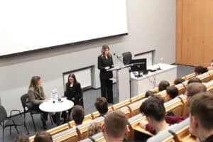 Aula uniwersytecka. Za białą mównicą stoi dr hab. Ewelina Podgajna, prof. UMCS, i przemawia do mikrofonu. Obok, przy małym okrągłym stoliku, siedzą Małgorzata Szumowska i Sylwia Kubica. Na pierwszym planie widać rzędy drewnianych ław z publicznością, głównie młodymi osobami. W tle znajduje się duży biały ekran projekcyjny.