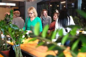 Grupa kobiet w różnym wieku rozmawia przy drewnianym blacie w jasnym wnętrzu. Jedna z nich, w zielonym swetrze i okularach, trzyma filiżankę. Na pierwszym planie rozmyte liście roślin