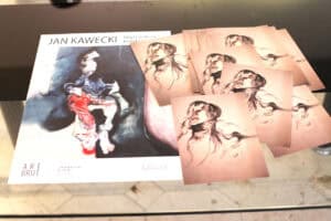 Materiały promocyjne wystawy artystycznej ułożone na szklanym blacie. Widoczny plakat z napisem „JAN KAWECKI” i logo galerii „ART BRUT” oraz ulotki z ekspresyjnymi rysunkami ludzkich postaci.