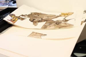 Stos luźnych arkuszy papieru leżących na stole. Na wierzchu widać rysunek przedstawiający fantastyczną istotę z dekoracyjnymi elementami w kolorze żółtym i niebieskim.