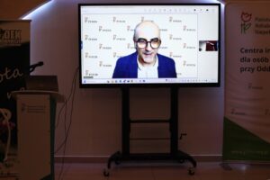 Na ekranie w sali konferencyjnej widoczny jest mężczyzna (Tomasz Wojakowski) w okularach, białej koszuli i niebieskiej marynarce, połączony zdalnie (online) jako prelegent. Ekran jest umieszczony obok mównicy PFRON oraz stojącego baneru PFRON
