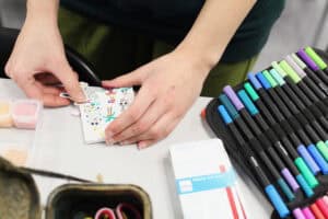 Zbliżenie na dłonie uczestniczki, która precyzyjnie nakleja drobne postacie i elementy na białą bazę pracy plastycznej. Obok leży otwarty piórnik z kolorowymi markerami i pudełko z koralikami.