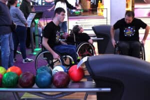 Stojak z wieloma kolorowymi kulami do kręgli (zielone, pomarańczowe, fioletowe i czerwona) na pierwszym planie. W tle widoczni są trzej uczestnicy zawodów na wózkach inwalidzkich, ubrani w czarne koszulki. Dwóch mężczyzn i kobieta oczekują na swoją kolej. Po lewej stronie stoją inni uczestnicy i kibice.