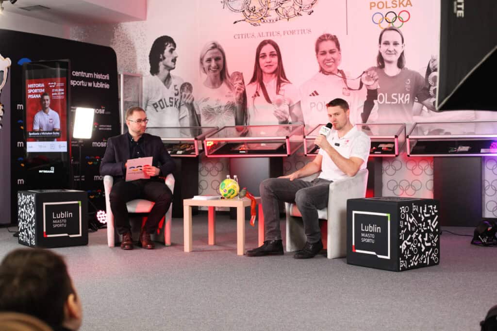 Od lewej siedzą prowadzący spotkanie oraz Michał Szyba. Sportowiec opiera jedną rękę na nodze a w drugiej trzyma Obok czarne pufy z napisem „Lublin Miasto Sportu”.