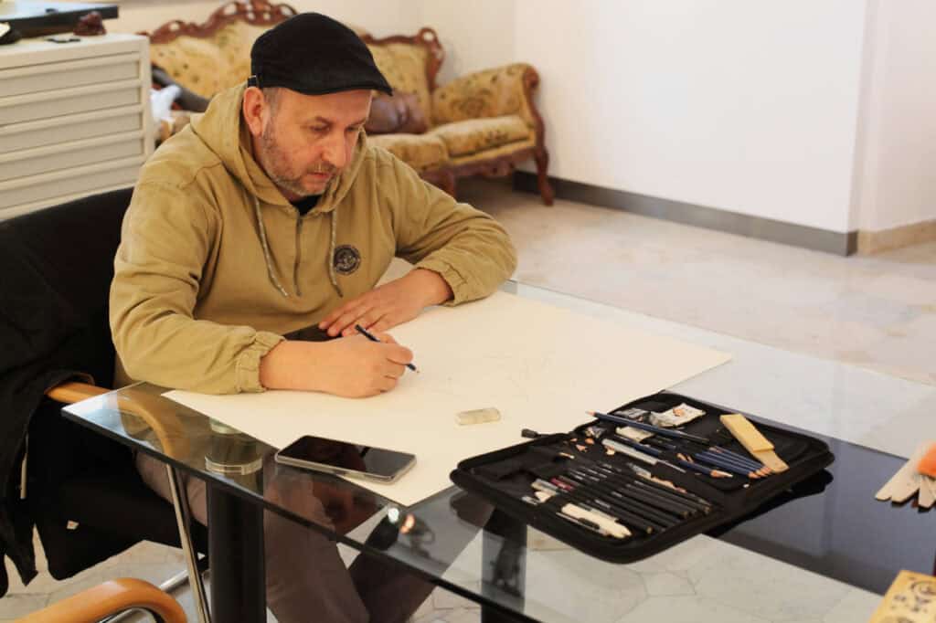 Mężczyzna w beżowej bluzie i czarnym kaszkiecie (Marcin Wrzal), siedzi przy szklanym stole i rysuje ołówkiem na dużym arkuszu papieru. Przed nim leży otwarte etui z profesjonalnymi ołówkami i akcesoriami kreślarskimi. W tle widoczny jest fragment sali galeryjnej z kanapą w stylu retro.