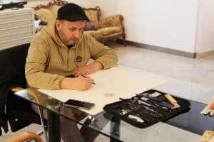 Mężczyzna w beżowej bluzie i czarnym kaszkiecie (Marcin Wrzal), siedzi przy szklanym stole i rysuje ołówkiem na dużym arkuszu papieru. Przed nim leży otwarte etui z profesjonalnymi ołówkami i akcesoriami kreślarskimi. W tle widoczny jest fragment sali galeryjnej z kanapą w stylu retro.