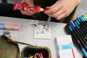Dłonie uczestniczki trzymają tubkę czerwonego kleju nad małą pracą plastyczną. Na białym podobraziu widoczne są kolorowe rysunki i naklejki. Wokół leżą organizery z koralikami i piórnik z pisakami.