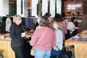 Rozmowy kuluarowe – Marta Rake trzyma kubek i rozmawia z innymi kobietami. W tle neon „SZWEDZKI STÓŁ”.