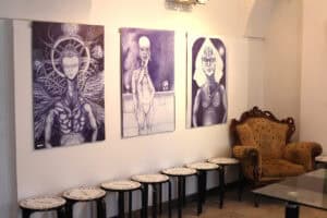 Trzy fioletowe grafiki wiszą na białej ścianie galerii. Pod nimi stoją taborety z napisem „TEATROTERAPIA LUBELSKA”