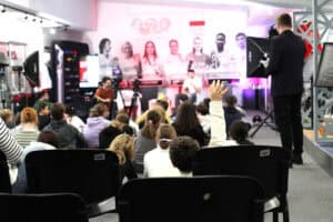 Ujęcie zza pleców publiczności siedzącej w rzędach krzeseł. Dziecko siedzące na widowni unosi rękę, w tle uczestnicy wywiadu na tle wystawy olimpijskiej.