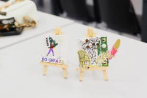 Dwie ukończone prace plastyczne stojące obok siebie na białym stole, każda na własnej drewnianej sztaludze. Lewa praca przedstawia postać z choinką na plecach i napis „ZIMA DO GWIAZD” ułożony z fioletowych liter. Prawa praca to kolaż z imieniem „KAMIL”, grafiką fotela i piórkiem.