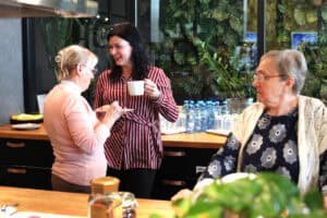 Kobiety rozmawiają przy bufecie. Jedna w pasiastej koszuli trzyma kubek, druga w różowym swetrze je ciasto.