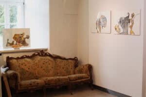 Od lewej: na parapecie na sztaludze stoi grafika, na sąsiedniej ścianie wiszą dwie prace w kolorach żółci, brązu i czerni. Poniżej stoi szeroka, bogato zdobiona kanapa w stylu retro.