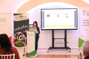 Kobieta w okularach prezentuje działalność "Powiatowego Zakładu Aktywności Zawodowej w Łęcznej" ze szczegółami dotyczącymi wyrobów i usług. Mównica PFRON stoi obok banerów: PFRON i Ośrodek Szkolno-Wychowawczy. Na lewo od mównicy stoi biały, wiklinowy kosz z ciemnoróżowymi orchideami