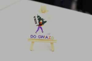 Praca plastyczna na małej sztaludze. Jest to miniaturowy kolaż na białym tle, przedstawiający postać w mikołajowej czapce niosącą dużą, zieloną choinkę. W lewym górnym rogu znajduje się pionowy napis „ZIMA”, a u dołu napis „DO GWIAZD” wykonany z wypukłych, fioletowych i zielonych liter. W prawym górnym rogu naklejono perłowe, wypukłe dżety imitujące słońce lub płatki śniegu.