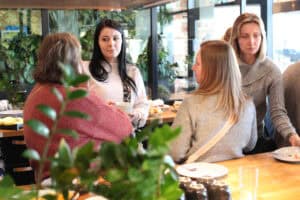 Cztery uczestniczki rozmawiają w grupie. Kobieta z długimi czarnymi włosami trzyma biały kubek. W tle przeszklona ściana i rośliny.